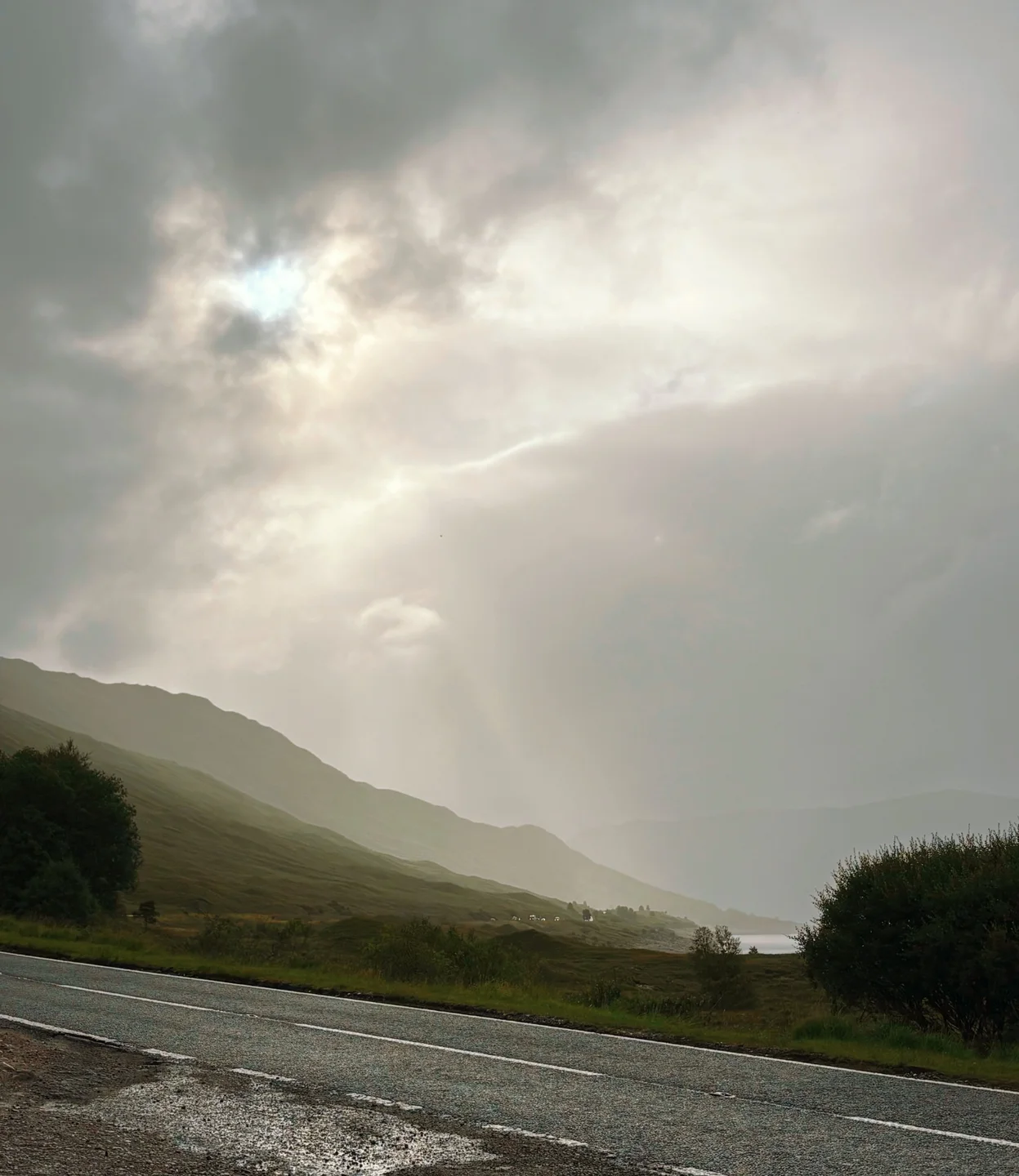 Sunlight breaking through heavy cloud above a wet roadside and distant hills.